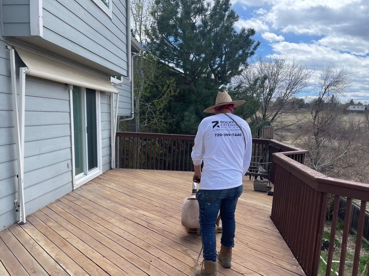 A crew member sanding a wood deck behind a gray-blue house