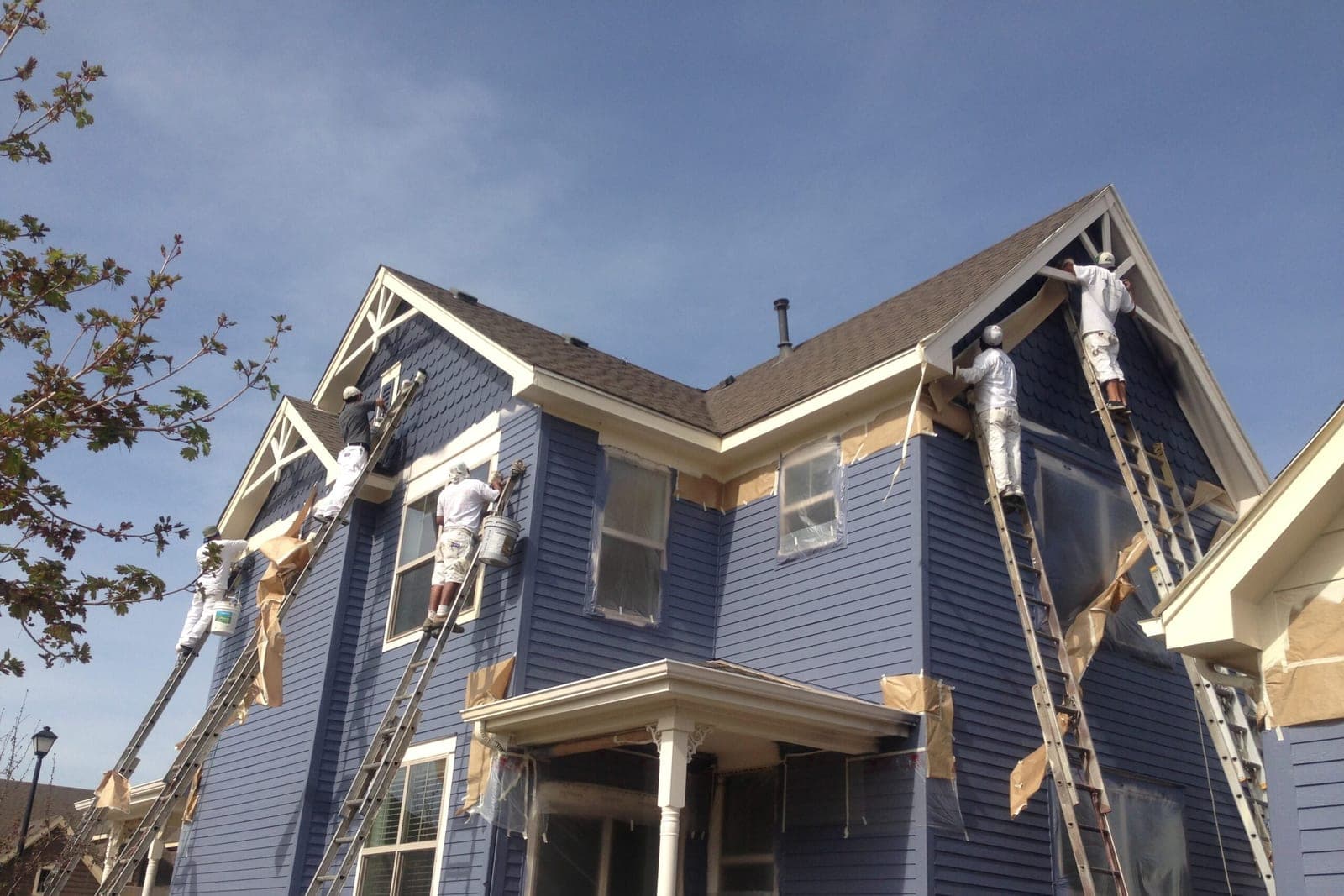 Four painters on ladders painting a blue Victorian-style house with cream trim